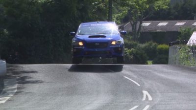 Professional rally driver Mark Higgins drives a blue Subaru WRX STI down a road on the Isle of Man.