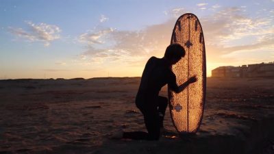 A man shaping a surfboard on the beach at sunset.