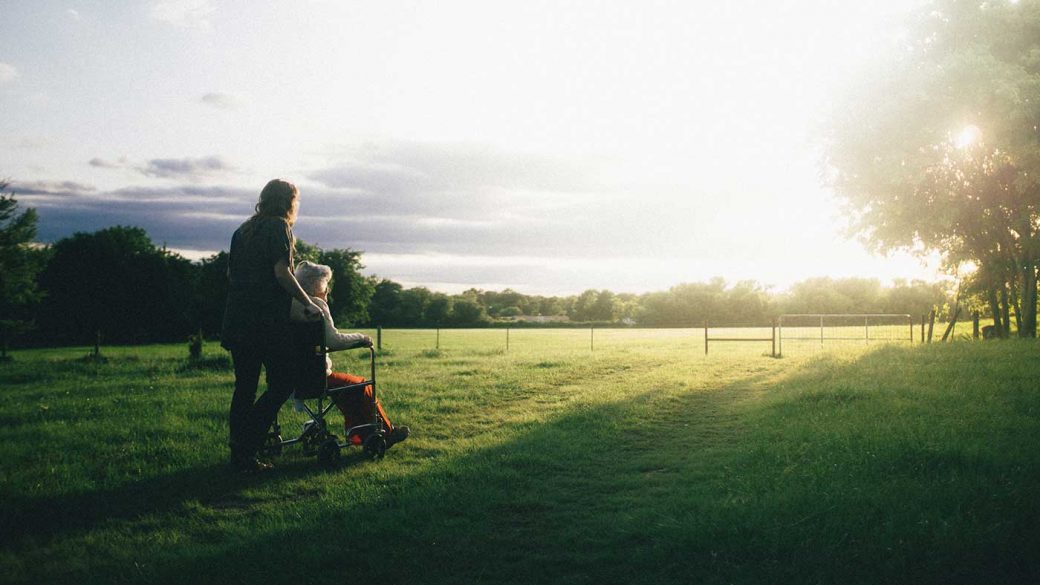 Elderly woman in a wheelchair being pushed by a carer through a field