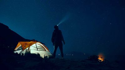 Man standing beside a tent, wearing a headlamp and looking up at a starry night sky