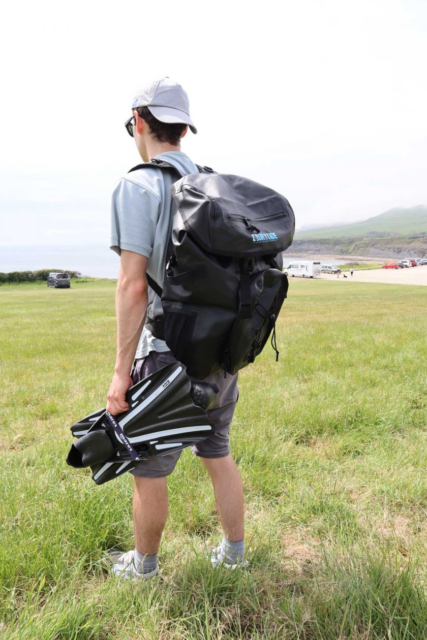 Looking to the left and carrying the DryTide 50L waterproof backpack whilst holding a pair of flippers