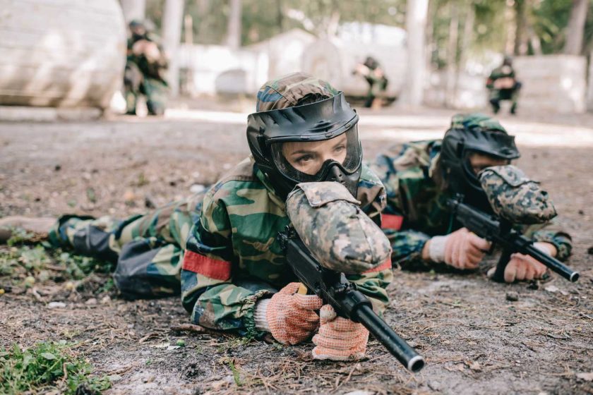 Two people playing paintball in a prone position wearing goggles and other paintball safety equipment