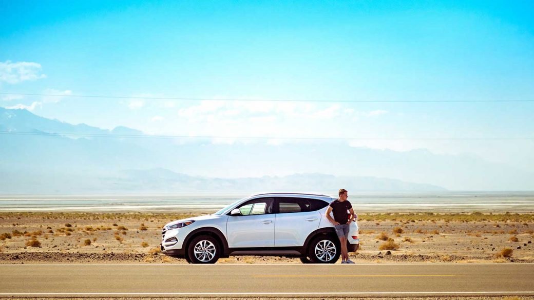 Man leaning against broken down white SUV on desert road