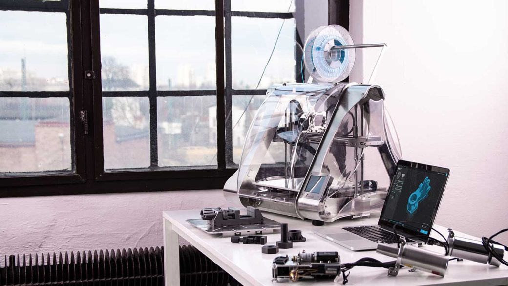 A 3D printer connected to a laptop on a white desk