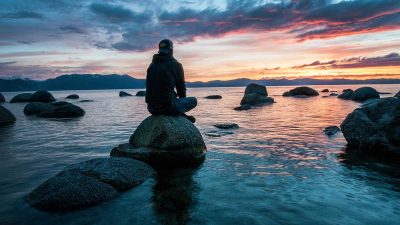 Man meditating on a rock in shallow water at sunset