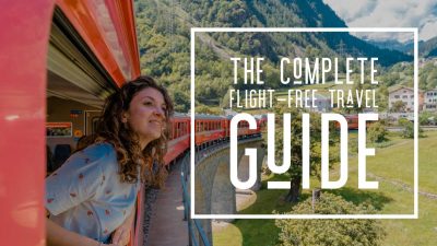 Woman travelling through the Swiss Alps in a red train, looking out of a window at the words ‘The complete flight-free travel guide’