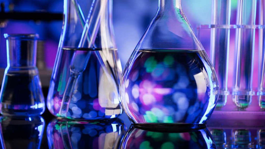Transparent laboratory flasks, filled with liquids, are illuminated by blue and purple lights, with a blurry colourful background.
