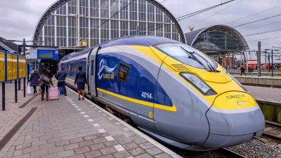 A Eurostar train is at the station platform with passengers eagerly boarding for a direct train journey from Amsterdam to London. The station boasts a large arched glass roof, adding a touch of grandeur to the bustling scene.
