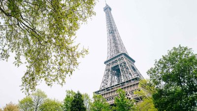 The Eiffel Tower in Paris is seen from below, framed by green leafy trees under a bright, slightly overcast sky—a must-see for first-timers exploring the city.