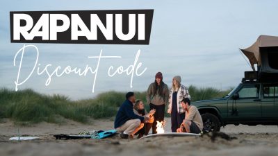 A group of people wearing organic cotton clothing sit around a beach campfire near a vehicle with a rooftop tent. Large text reads "Rapanui clothing 15% OFF discount code.